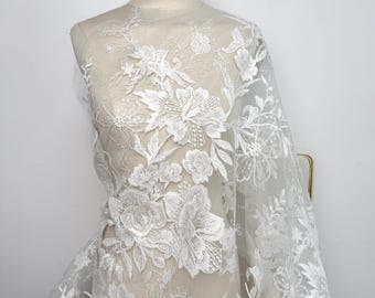 May include: White floral lace fabric draped over a mannequin. The lace features detailed, raised floral embroidery with a variety of flower and leaf designs. The fabric is sheer, allowing the mannequin to be partially visible.