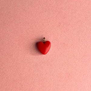 Puede incluir: Un pequeño colgante rojo brillante en forma de corazón con un lazo dorado para colgar. El colgante está centrado en un fondo rosa claro texturizado, creando una estética simple y romántica. Ideal para la fabricación de joyas.