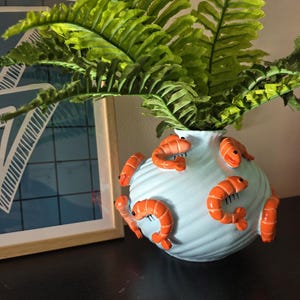 May include: A light blue ceramic vase decorated with orange shrimp. The vase holds green fern-like plants. The vase has a textured, wavy design. A framed picture is in the background.