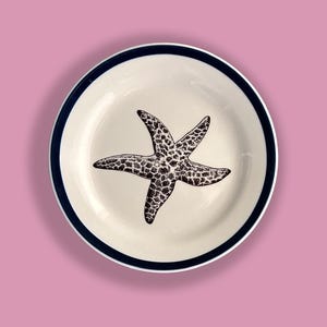 May include: A white ceramic plate with a blue rim features a black and white starfish design.