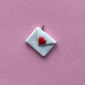 以下が含まれることがあります： ピンク色の背景に、赤いハートのアクセントが付いた白い封筒のペンダント。ペンダントには、吊り下げ用の金色のループが付いています。封筒は正方形で、滑らかで光沢のある素材でできているようです。