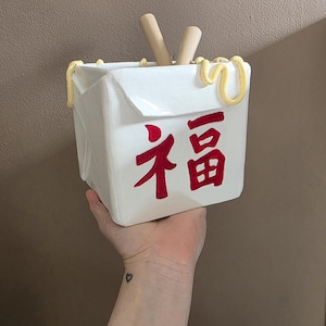 Könnte beinhalten: Ein weißer Keramik-Imbissbehälter mit roten chinesischen Schriftzeichen und gelben Akzenten. Zwei Holzstäbchen befinden sich im Inneren des Behälters. Der Behälter wird von einer Hand gehalten.
