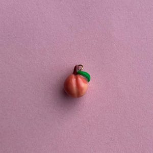 Puede incluir: Un pequeño dije de melocotón con una hoja verde y un lazo dorado. El melocotón es una mezcla de tonos rosados y anaranjados, con sutiles estrías. El dije es probablemente para joyería o manualidades.