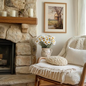 May include: A white crocheted armchair with a cream throw blanket and a white pillow. A bouquet of white daisies in a cream ceramic vase sits on a wooden table next to the chair. A framed picture of a tree with a birdhouse hangs on the wall behind the chair.