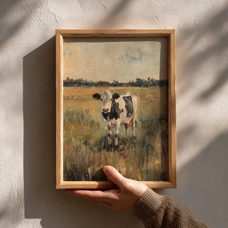 Farmhouse cow print being held by hand in sunlight, rustic wooden frame casting shadow on cream wall