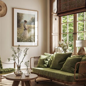 May include: A green velvet sofa with a woven wood frame and a wooden coffee table with a vase of white flowers. A framed painting of a woman in a white dress standing in a field of flowers hangs on the wall.