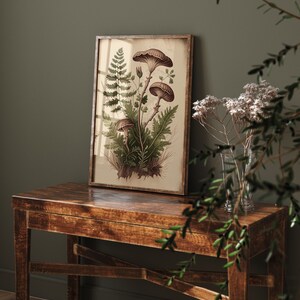 May include: A framed botanical print featuring a cluster of brown mushrooms and green ferns on a cream background. The print is displayed on a dark brown wooden table.