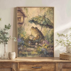 Puede incluir: Impresión artística enmarcada que representa una rana con gafas, sosteniendo una taza de té bajo un paraguas. La rana está rodeada de flores y vegetación, con una pequeña casa al fondo. La obra tiene un estilo vintage y caprichoso.