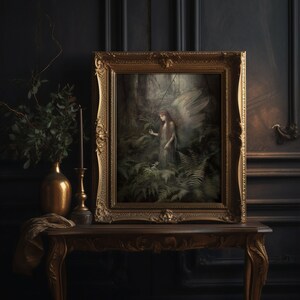 May include: A framed artwork featuring a forest scene with an angel figure, displayed on a wooden table. The artwork is in a gold ornate frame. A gold vase with greenery and a candle holder are on the table, set against a dark wall.
