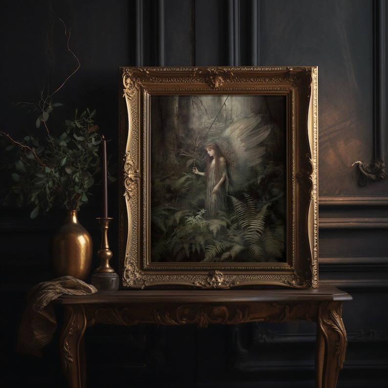 May include: A framed artwork featuring a forest scene with an angel figure, displayed on a wooden table. The artwork is in a gold ornate frame. A gold vase with greenery and a candle holder are on the table, set against a dark wall.