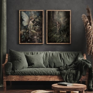 May include: Two framed art prints depicting ethereal fairies in a forest setting. The artwork features soft colors, with a focus on nature and fantasy themes. The frames are a warm wood tone, complementing the dark wall and green sofa.