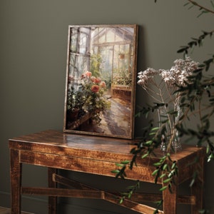 May include: A framed painting of a greenhouse with flowers and a stone path. The painting is on a wooden table with a vase of dried flowers.