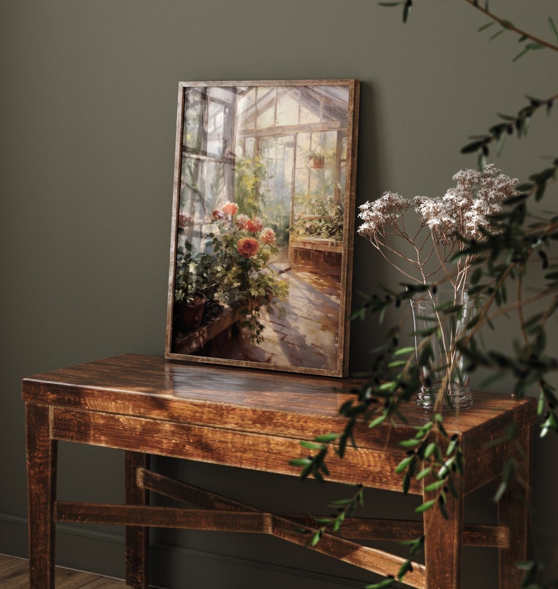 May include: A framed painting of a greenhouse with flowers and a stone path. The painting is on a wooden table with a vase of dried flowers.