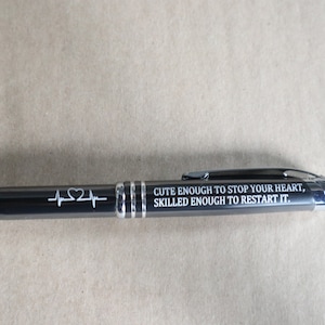 May include: A black pen with a silver clip and a stylus tip. The pen has a heartbeat and heart design engraved on it and the text "Cute enough to stop your heart, skilled enough to restart it."