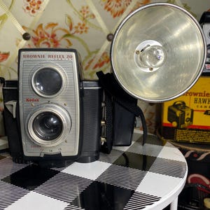 May include: A vintage Kodak Brownie Reflex 20 camera with a flash attachment. The camera is black and silver, with the text "Brownie Reflex 20" on the front. The flash has a large, silver reflector. The camera sits on a black and white checkered table.