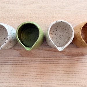 May include: Four ceramic creamers in a row, each with a unique color and speckled design. The colors include white, green, white, and tan. Each creamer has a spout for easy pouring. The creamers are arranged on a light wood surface.