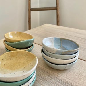 May include: A stack of small, handmade ceramic bowls in yellow, green, and blue speckled glazes. The bowls are stacked on a light wood surface.