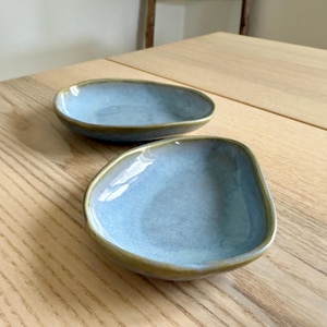 May include: Two small, light blue ceramic dishes with irregular shapes and a golden-brown rim. The dishes are placed on a light-colored wooden surface, creating a simple and elegant aesthetic. The dishes are likely for serving small portions.