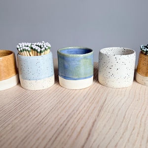 May include: Five ceramic match holders in various colors, including speckled white, blue, and tan. Some holders contain matches with green or white tips. Each holder has a white base and a cylindrical shape.