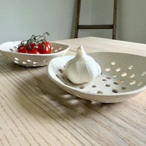 May include: Two off-white ceramic bowls with a speckled finish and numerous circular cutouts along the sides and base. One bowl contains a cluster of red cherry tomatoes on the vine, while the other holds a single bulb of white garlic.