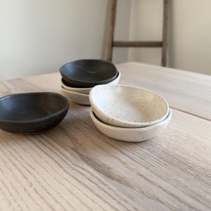 May include: A set of small, round ceramic bowls. The bowls are in two colors: black and white with speckles. The bowls are stacked on a light wood surface.