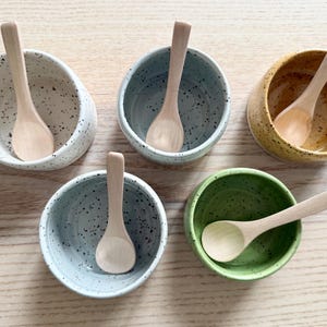 May include: A collection of five speckled ceramic bowls in white, blue, yellow, and green, each containing a small wooden spoon. The bowls are arranged on a light-colored wooden surface, showcasing a rustic, handcrafted aesthetic.