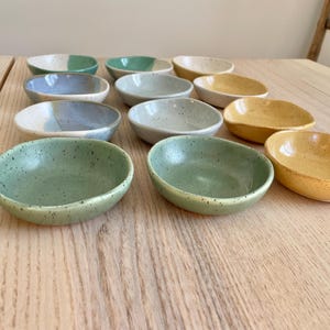 May include: A set of ten small, speckled ceramic bowls in various shades of green, blue, and yellow. The bowls are arranged in a staggered pattern on a wooden surface.