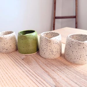 May include: Four ceramic vessels with speckled patterns. Two are off-white with black speckles, one is green, and the last is off-white with black speckles. The vessels are cylindrical with a slightly angled top, sitting on a light wood surface.