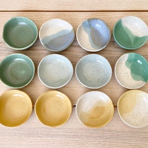 May include: Twelve small ceramic bowls in various colors and patterns. The bowls are arranged in three rows of four. The bowls are all different colors and patterns, including green, blue, yellow, and white.