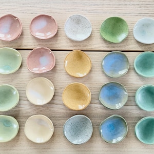 May include: An overhead shot displays a collection of twenty small, handmade ceramic dishes. The dishes are round with slightly irregular edges and come in a variety of pastel colors, including pink, green, blue, and yellow. The dishes are arranged on a light-colored wooden surface.
