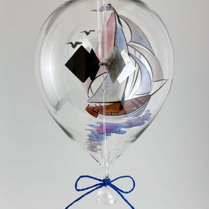 Könnte beinhalten: Ein klarer Glas-Windspiel mit einem Segelboot-Design im Inneren. Das Segelboot hat einen braunen Rumpf, weiße Segel und einen rosa und blauen Himmel. Das Windspiel hat eine blaue Schnur und eine dekorative Schleife.