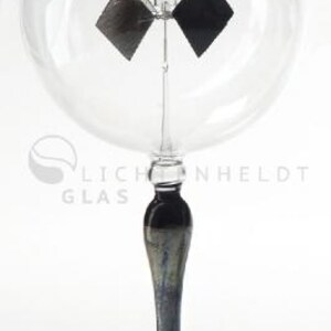 Puede incluir: Una esfera de vidrio con una hélice negra y plateada en su interior, montada sobre un soporte de vidrio negro e iridiscente. La esfera está etiquetada con "Licht und Heldt Glas".
