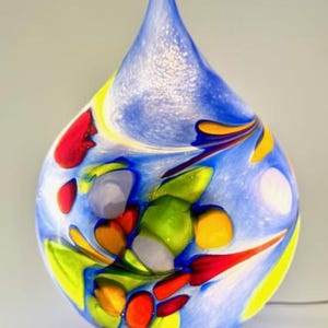 Può includere: Lampada decorativa in vetro a forma di goccia. La lampada presenta uno sfondo blu con accenti colorati di rosso, giallo, verde e arancione. Poggia su una base rotonda in legno e ha una parte superiore appuntita.
