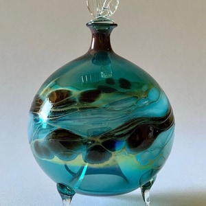 Puede incluir: Un frasco de perfume esférico de vidrio en un tono azul cerceta vibrante, con patrones arremolinados de color marrón oscuro y dorado en todo el cuerpo. El recipiente se apoya sobre tres pequeñas patas de vidrio transparente y tiene un tapón.