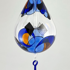 Peut inclure: Une décoration en verre en forme de larme avec un motif abstrait bleu, orange et noir. La décoration a un corps en verre transparent et une larme en verre bleu suspendue au bas.