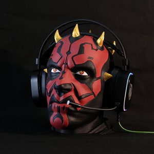May include: A black gaming headset with red and gold accents. The headset has a microphone and is worn by a red and gold Darth Maul mask.