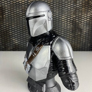 May include: A silver and black 3D printed bust of a Mandalorian character from the Star Wars franchise. The bust features a detailed helmet and armor, including a jetpack and a blaster rifle.