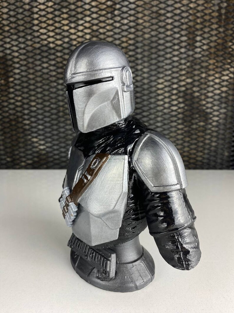 May include: A silver and black 3D printed bust of a Mandalorian character from the Star Wars franchise. The bust features a detailed helmet and armor, including a jetpack and a blaster rifle.