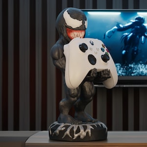 May include: A black and white Venom figurine holding a white Xbox controller. The figurine has a detailed design, with a menacing grin and a textured base. The controller has multiple buttons and joysticks.