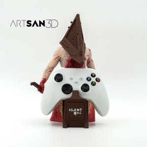 以下が含まれることがあります： 白いビデオゲームコントローラーは、サイレントヒルビデオゲームシリーズのキャラクターの3Dプリントフィギュアによって保持されています。フィギュアは茶色のピラミッド型のヘルメットをかぶり、血まみれの胴体を持っています。コントローラースタンドは茶色で、「サイレントヒル」という文字が印刷されています。