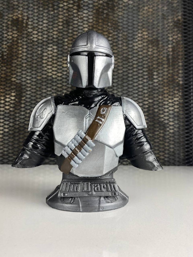 May include: A silver and black Mandalorian bust with a brown bandolier and the text "The Mandalorian" on the base.