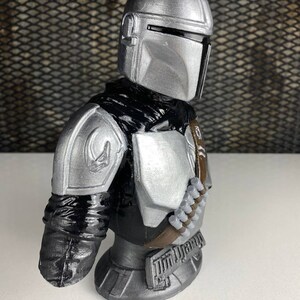 May include: A silver and black Mandalorian bust figurine. The bust is detailed with a helmet, armor, and a bandolier.