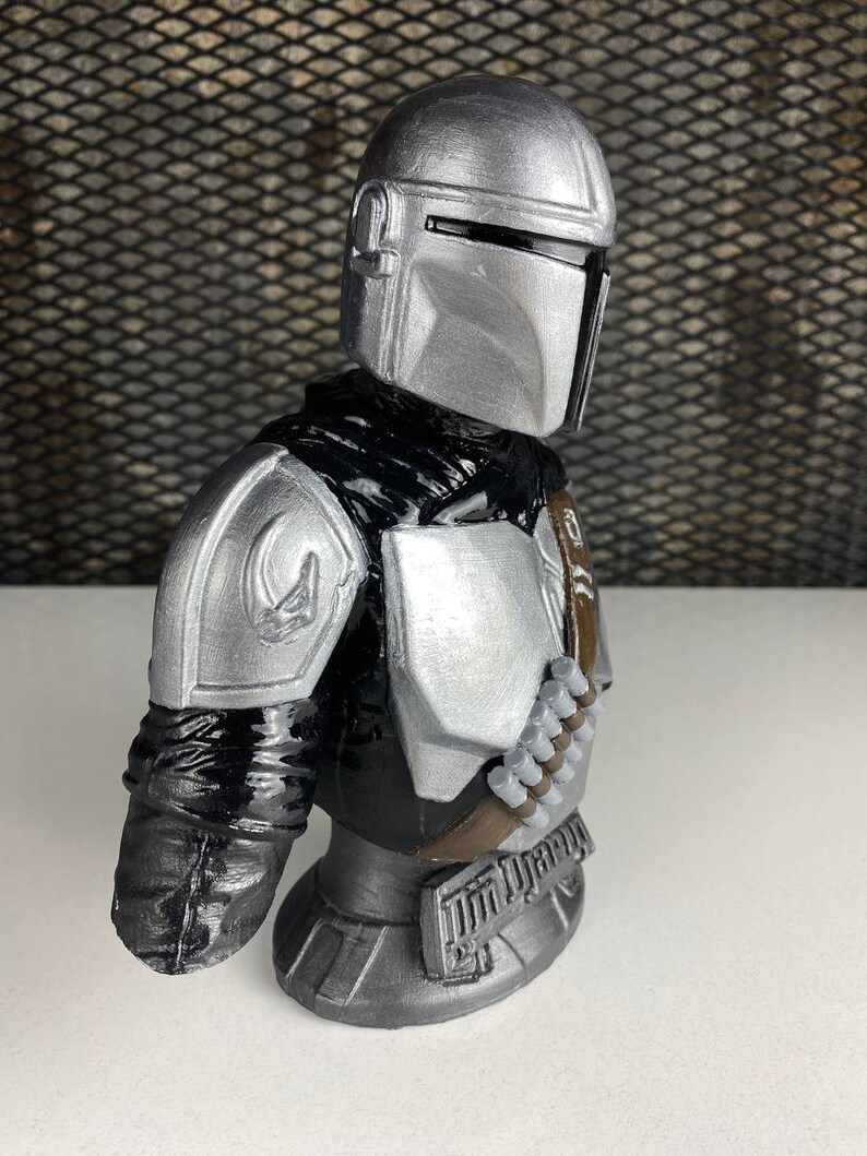 May include: A silver and black Mandalorian bust figurine. The bust is detailed with a helmet, armor, and a bandolier.