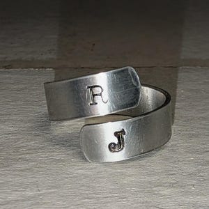 May include: A silver-colored wrap ring with the letters "R" and "J" stamped on the band. The ring has a simple, minimalist design and is likely made of a metal like aluminum or stainless steel.