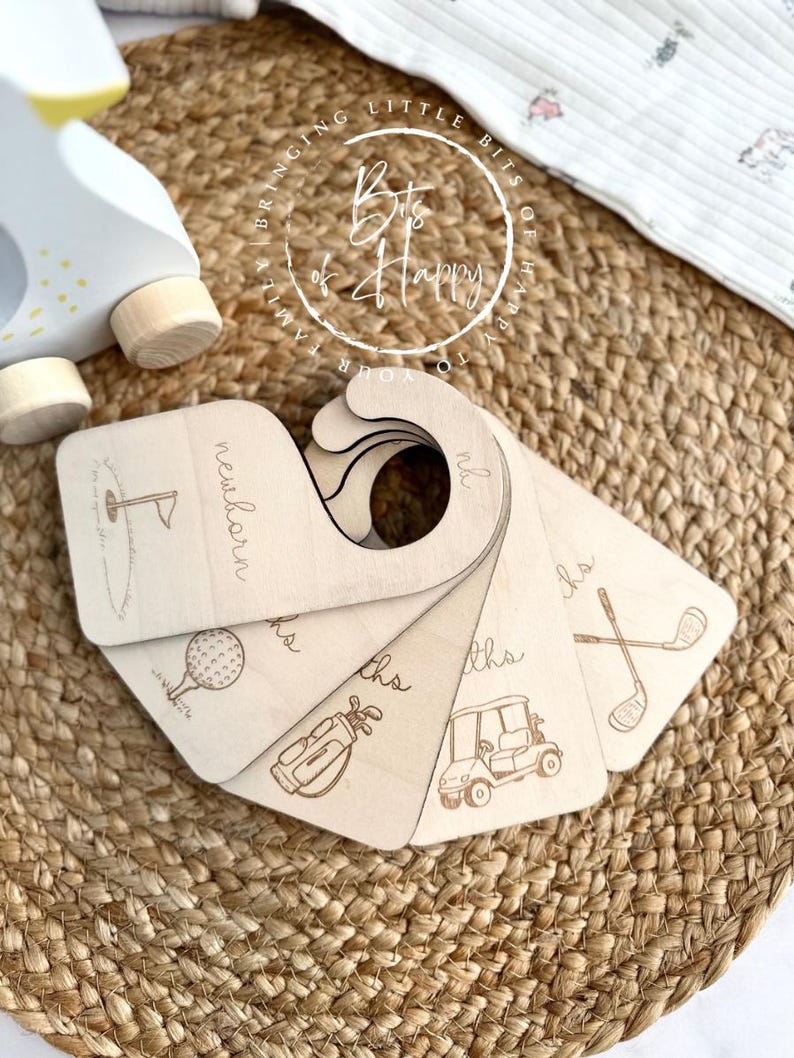 May include: Wooden baby milestone markers with golf-themed illustrations. Each marker is engraved with a different golf-related image and the words "newborn" and "months". The markers are arranged in a fan shape.
