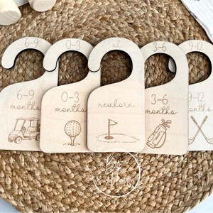 May include: Five wooden baby clothes hangers with golf-themed illustrations. Each hanger is labeled with a size range: 6-9 months, 0-3 months, newborn, 3-6 months, and 9-12 months. The hangers are made of light-colored wood.