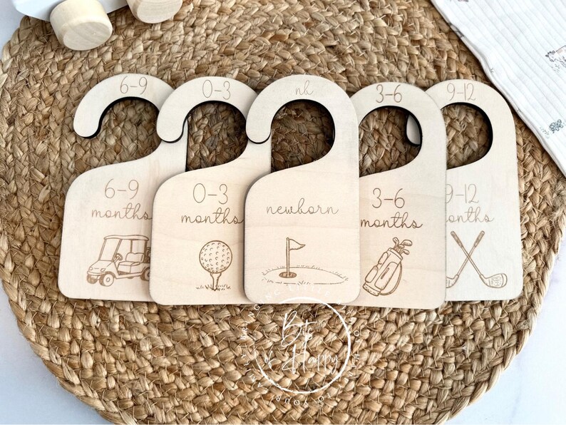 May include: Five wooden baby clothes hangers with golf-themed illustrations. Each hanger is labeled with a size range: 6-9 months, 0-3 months, newborn, 3-6 months, and 9-12 months. The hangers are made of light-colored wood.