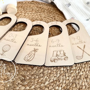 May include: Wooden baby clothes hangers with golf-themed illustrations and age ranges: newborn, 0-3 months, 3-6 months, 6-9 months, and 9-12 months. The hangers are arranged on a woven mat.