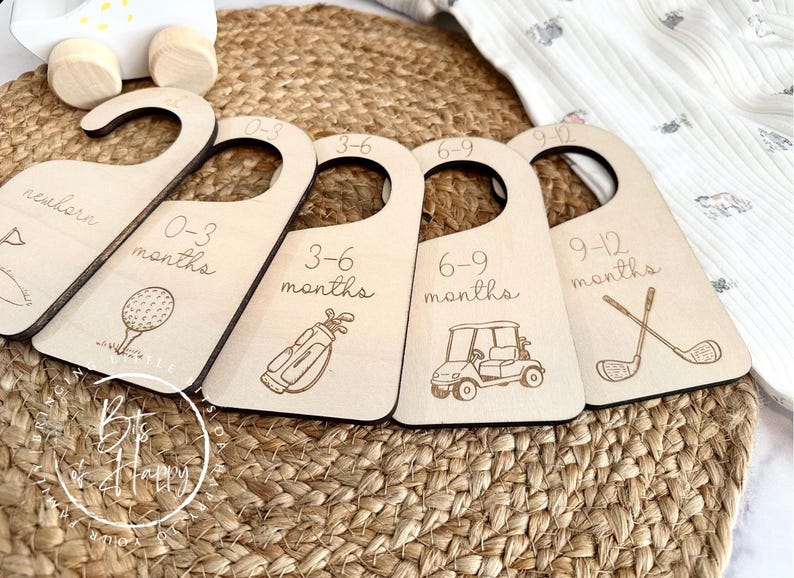 May include: Wooden baby clothes hangers with golf-themed illustrations and age ranges: newborn, 0-3 months, 3-6 months, 6-9 months, and 9-12 months. The hangers are arranged on a woven mat.