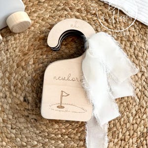 May include: A wooden baby clothes hanger with the word "newborn" and a golf flag design. The hanger has the initials "nh" on the top and is adorned with a white ribbon. The background is a woven, natural-colored material.